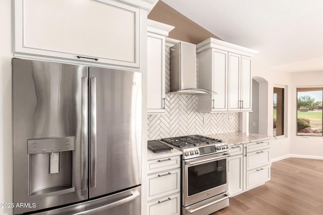 kitchen with light wood-style flooring, appliances with stainless steel finishes, backsplash, light stone countertops, and wall chimney exhaust hood