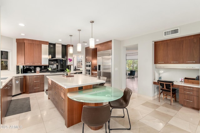 kitchen with a kitchen island, built in desk, hanging light fixtures, stainless steel appliances, and wall chimney exhaust hood