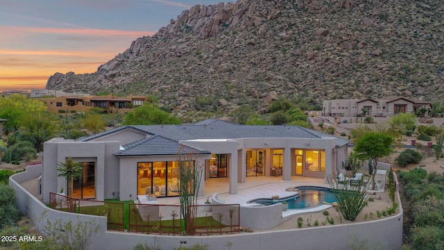 back of property at dusk featuring a mountain view, fence, a patio, stucco siding, and an in ground hot tub