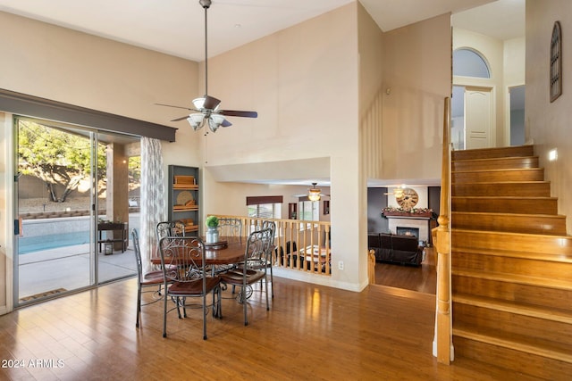 dining room with a warm lit fireplace, ceiling fan, a towering ceiling, and wood finished floors