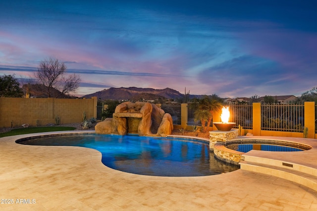 pool at dusk with a patio area, a pool with connected hot tub, a mountain view, and fence