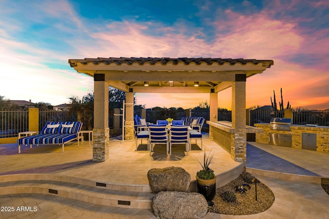 patio terrace at dusk featuring exterior kitchen, area for grilling, outdoor dining area, and fence