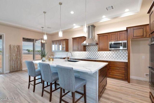 kitchen featuring decorative light fixtures, wall chimney range hood, sink, a kitchen island with sink, and appliances with stainless steel finishes