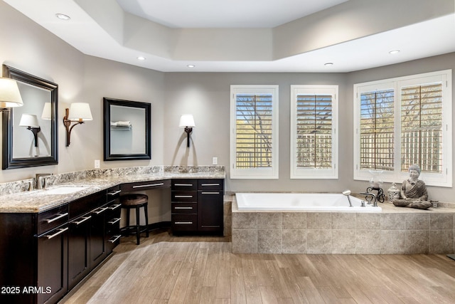 bathroom featuring a raised ceiling, wood-type flooring, a relaxing tiled tub, and vanity