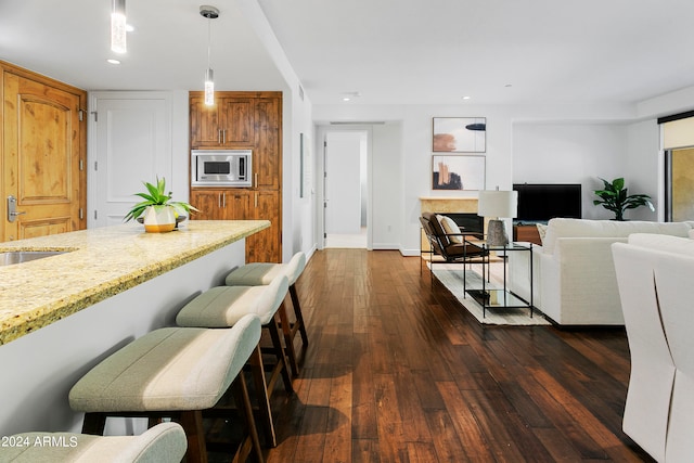 interior space featuring dark wood-type flooring, stainless steel microwave, hanging light fixtures, a kitchen breakfast bar, and light stone countertops