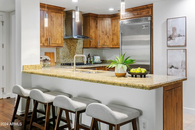 kitchen with a breakfast bar area, kitchen peninsula, built in refrigerator, wall chimney exhaust hood, and decorative light fixtures