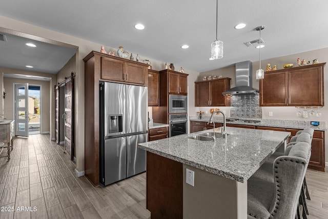 kitchen featuring appliances with stainless steel finishes, a kitchen island with sink, sink, wall chimney range hood, and hanging light fixtures