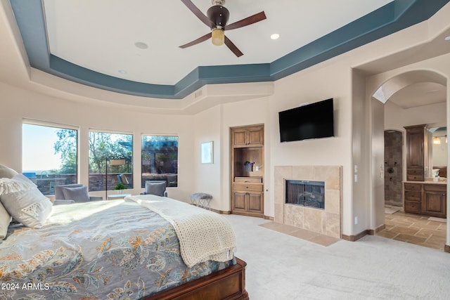 carpeted bedroom featuring ceiling fan, connected bathroom, a tiled fireplace, and a raised ceiling