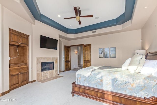 bedroom featuring a tiled fireplace, ceiling fan, a raised ceiling, and light carpet