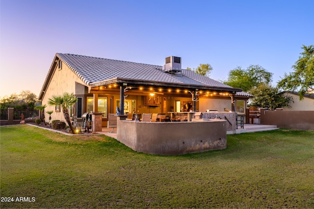 back house at dusk with a lawn, an outdoor kitchen, exterior bar, and central AC