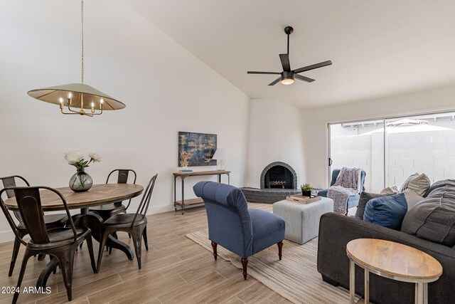 living area featuring baseboards, high vaulted ceiling, light wood-style flooring, a brick fireplace, and ceiling fan with notable chandelier