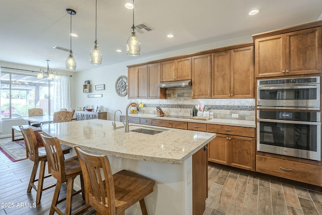 kitchen featuring stainless steel double oven, under cabinet range hood, a sink, light wood finished floors, and tasteful backsplash