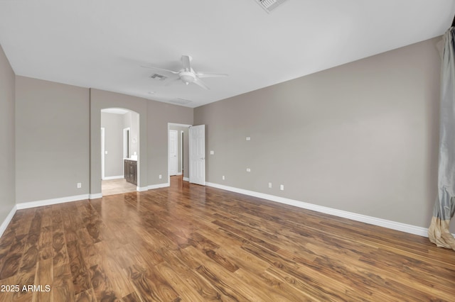 unfurnished bedroom featuring arched walkways, wood finished floors, baseboards, ensuite bathroom, and ceiling fan