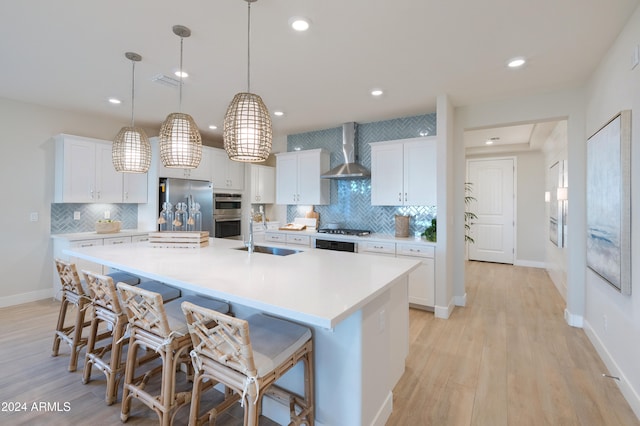 kitchen with appliances with stainless steel finishes, wall chimney exhaust hood, pendant lighting, a large island with sink, and white cabinetry