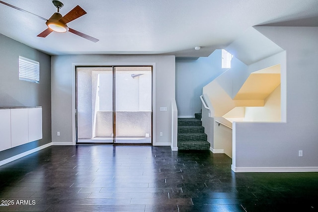 unfurnished living room featuring a ceiling fan, stairs, baseboards, and dark wood-type flooring