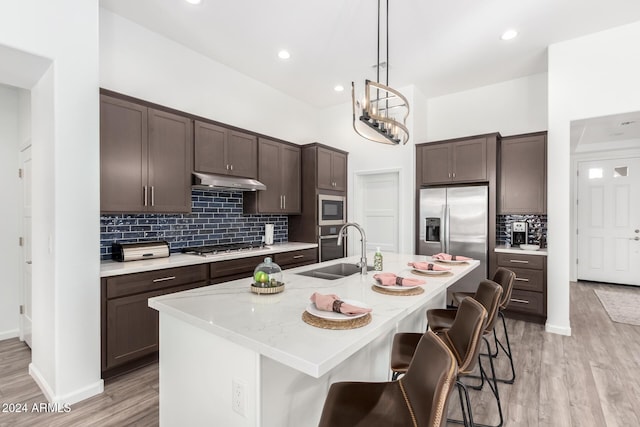 kitchen with dark brown cabinets, stainless steel appliances, a kitchen island with sink, sink, and light hardwood / wood-style flooring