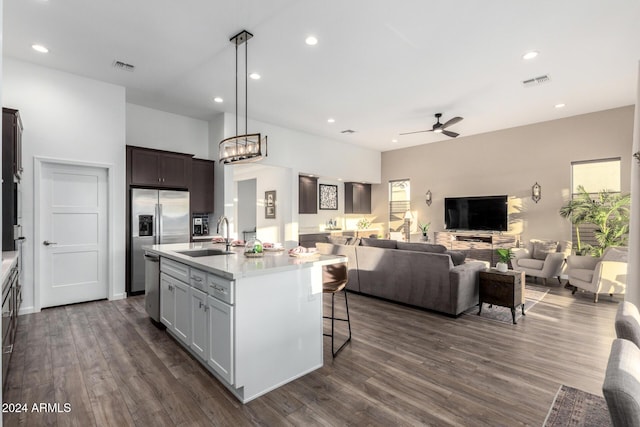 kitchen with ceiling fan with notable chandelier, stainless steel appliances, dark wood-type flooring, sink, and an island with sink