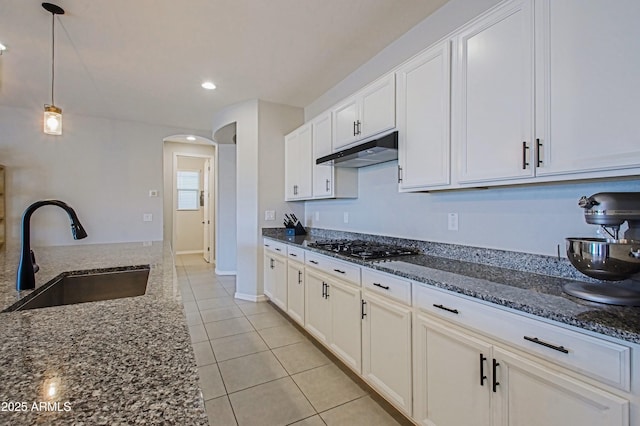 kitchen featuring arched walkways, stainless steel gas cooktop, a sink, dark stone countertops, and under cabinet range hood