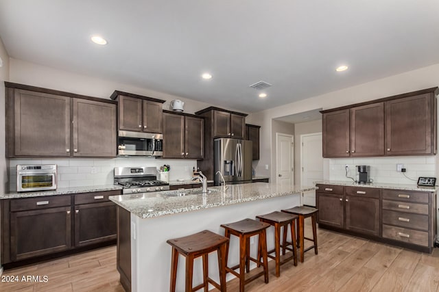 kitchen featuring a kitchen island with sink, light hardwood / wood-style floors, appliances with stainless steel finishes, decorative backsplash, and a kitchen breakfast bar