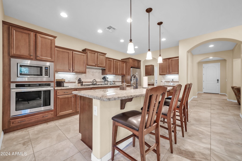 kitchen with a kitchen bar, stainless steel appliances, pendant lighting, light stone counters, and a kitchen island with sink