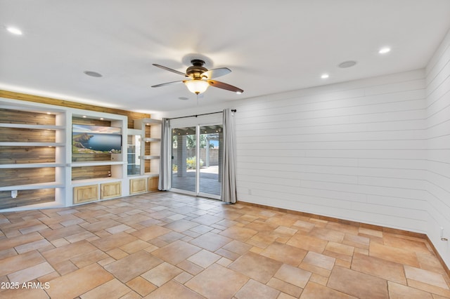 empty room with built in shelves, a sunroom, ceiling fan, and wood walls