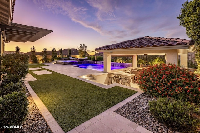 pool at dusk featuring a gazebo, exterior kitchen, pool water feature, a patio area, and a lawn