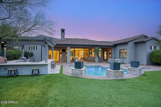 back house at dusk featuring exterior bar, exterior kitchen, a patio area, and a lawn