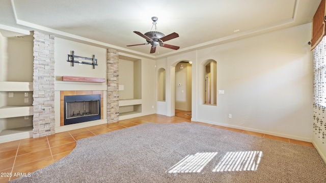 unfurnished living room with arched walkways, a stone fireplace, tile patterned flooring, a ceiling fan, and a tray ceiling