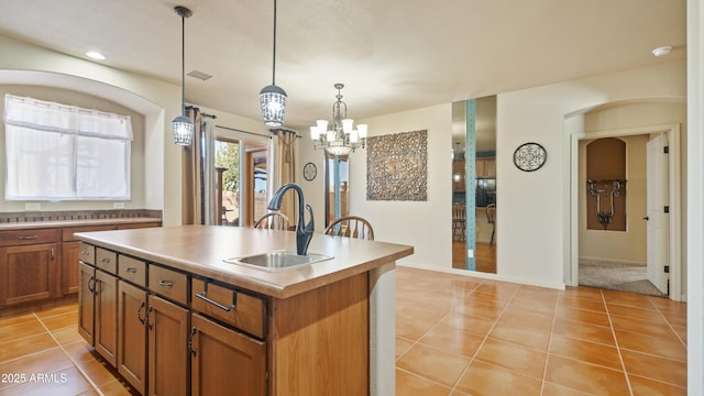 kitchen with light tile patterned floors, a sink, brown cabinetry, decorative light fixtures, and an island with sink