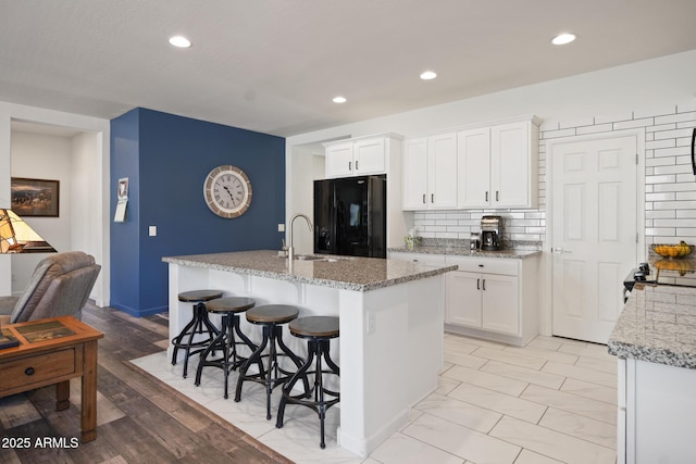 kitchen featuring black refrigerator, white cabinets, a kitchen island with sink, and light stone counters