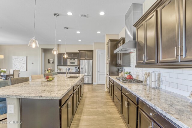 kitchen featuring hanging light fixtures, stainless steel appliances, a kitchen bar, a kitchen island with sink, and wall chimney range hood