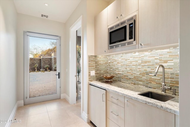 kitchen featuring light brown cabinets, stainless steel microwave, backsplash, sink, and light stone counters