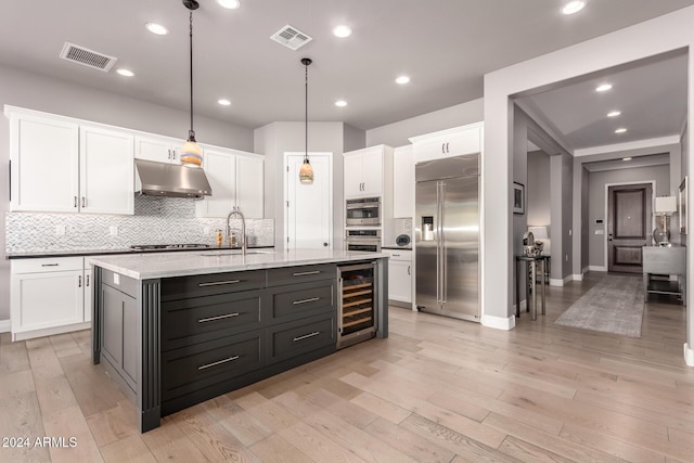 kitchen featuring a center island with sink, light wood-type flooring, appliances with stainless steel finishes, decorative light fixtures, and beverage cooler