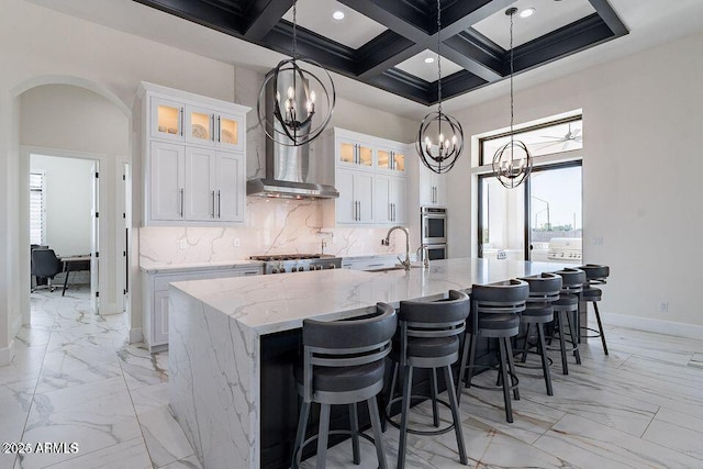 kitchen featuring light stone counters, glass insert cabinets, coffered ceiling, white cabinets, and a center island with sink