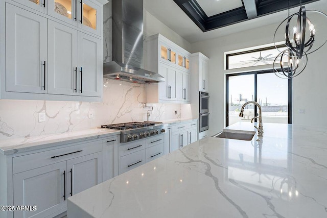 kitchen with appliances with stainless steel finishes, glass insert cabinets, light stone countertops, wall chimney range hood, and a sink