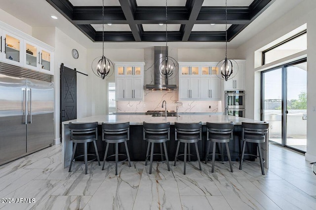 kitchen with a barn door, stainless steel appliances, white cabinets, wall chimney range hood, and glass insert cabinets