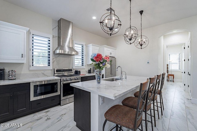 kitchen featuring appliances with stainless steel finishes, white cabinets, a kitchen island with sink, and wall chimney range hood