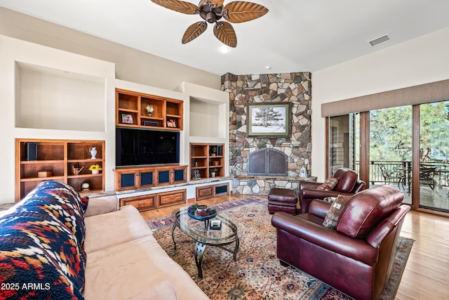 living room featuring ceiling fan, a stone fireplace, built in features, and light wood-type flooring