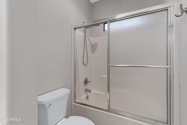 bathroom featuring toilet, a textured wall, and combined bath / shower with glass door