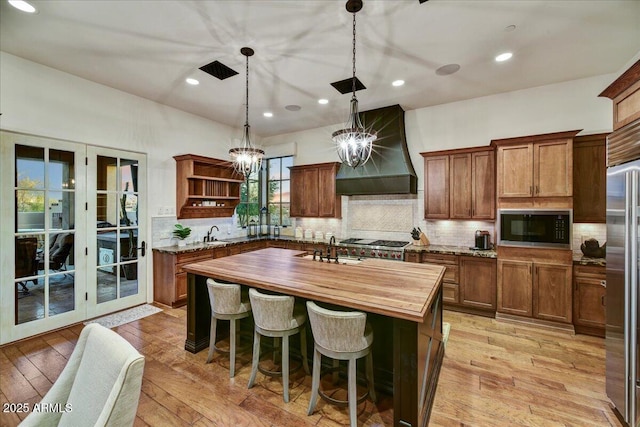 kitchen with custom exhaust hood, open shelves, a kitchen island with sink, built in appliances, and wood counters