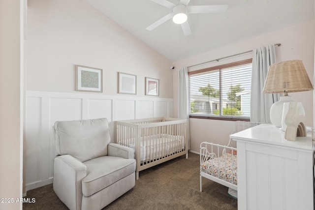 carpeted bedroom featuring vaulted ceiling, wainscoting, a crib, a decorative wall, and ceiling fan