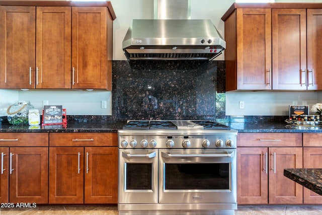 kitchen with dark stone counters, wall chimney range hood, brown cabinetry, and range with two ovens