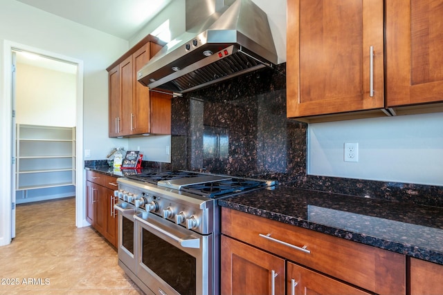 kitchen featuring range with two ovens, wall chimney range hood, decorative backsplash, dark stone counters, and brown cabinetry