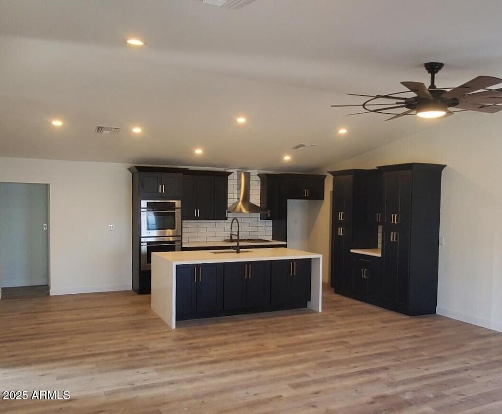 kitchen with tasteful backsplash, sink, a kitchen island with sink, double oven, and wall chimney exhaust hood