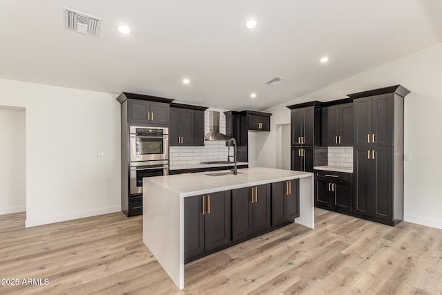 kitchen with lofted ceiling, decorative backsplash, an island with sink, stainless steel appliances, and wall chimney exhaust hood