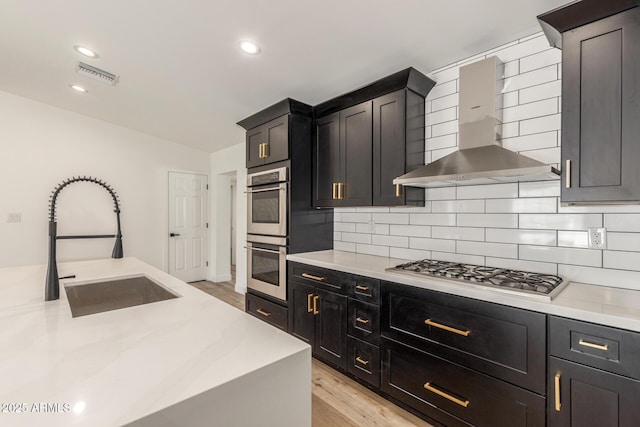 kitchen with stainless steel appliances, backsplash, light wood-type flooring, wall chimney range hood, and sink