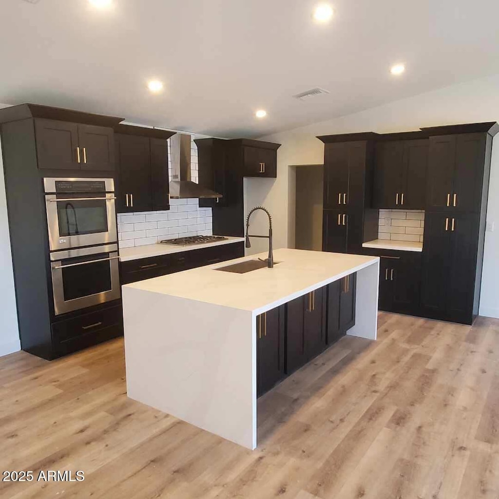 kitchen with sink, wall chimney range hood, a kitchen island with sink, and tasteful backsplash