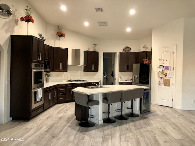 kitchen with stainless steel appliances, wall chimney range hood, light hardwood / wood-style floors, a kitchen island with sink, and dark brown cabinets