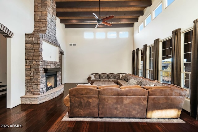 living room with ceiling fan, beam ceiling, a towering ceiling, wood-type flooring, and a stone fireplace