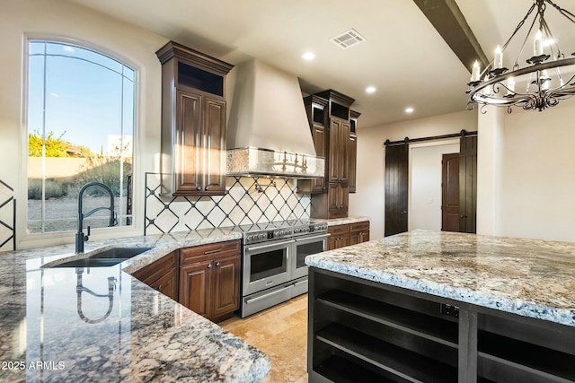 kitchen with sink, range with two ovens, light stone counters, a barn door, and custom range hood